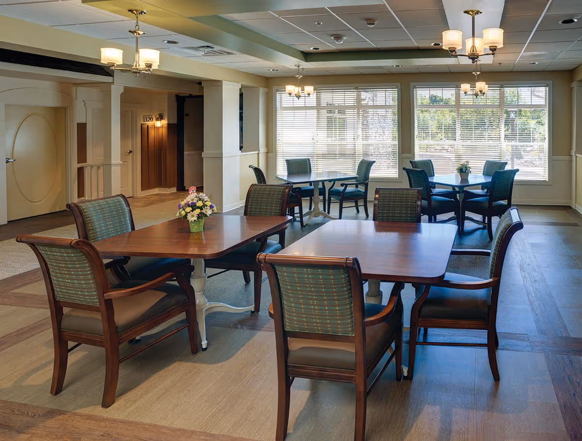 Bright common dining area with wooden tables, upholstered chairs, and large windows.