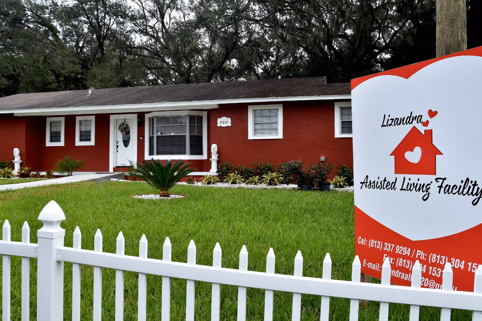 Front view of a red single-story assisted living facility with a white picket fence.