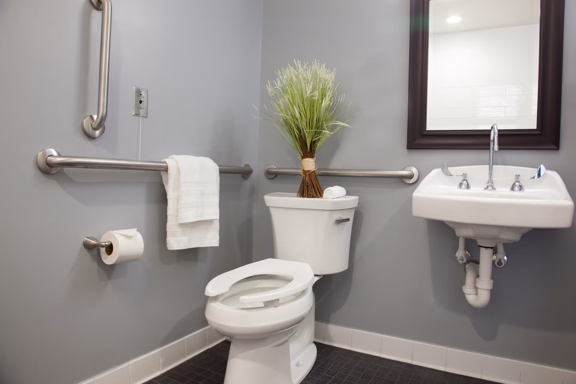 A clean, accessible bathroom with gray walls featuring a white toilet with an open seat, a white sink with a mirror above it, stainless steel grab bars on the walls, a towel hanging on one grab bar, a roll of toilet paper on a holder, and a decorative plant placed on the toilet tank.
