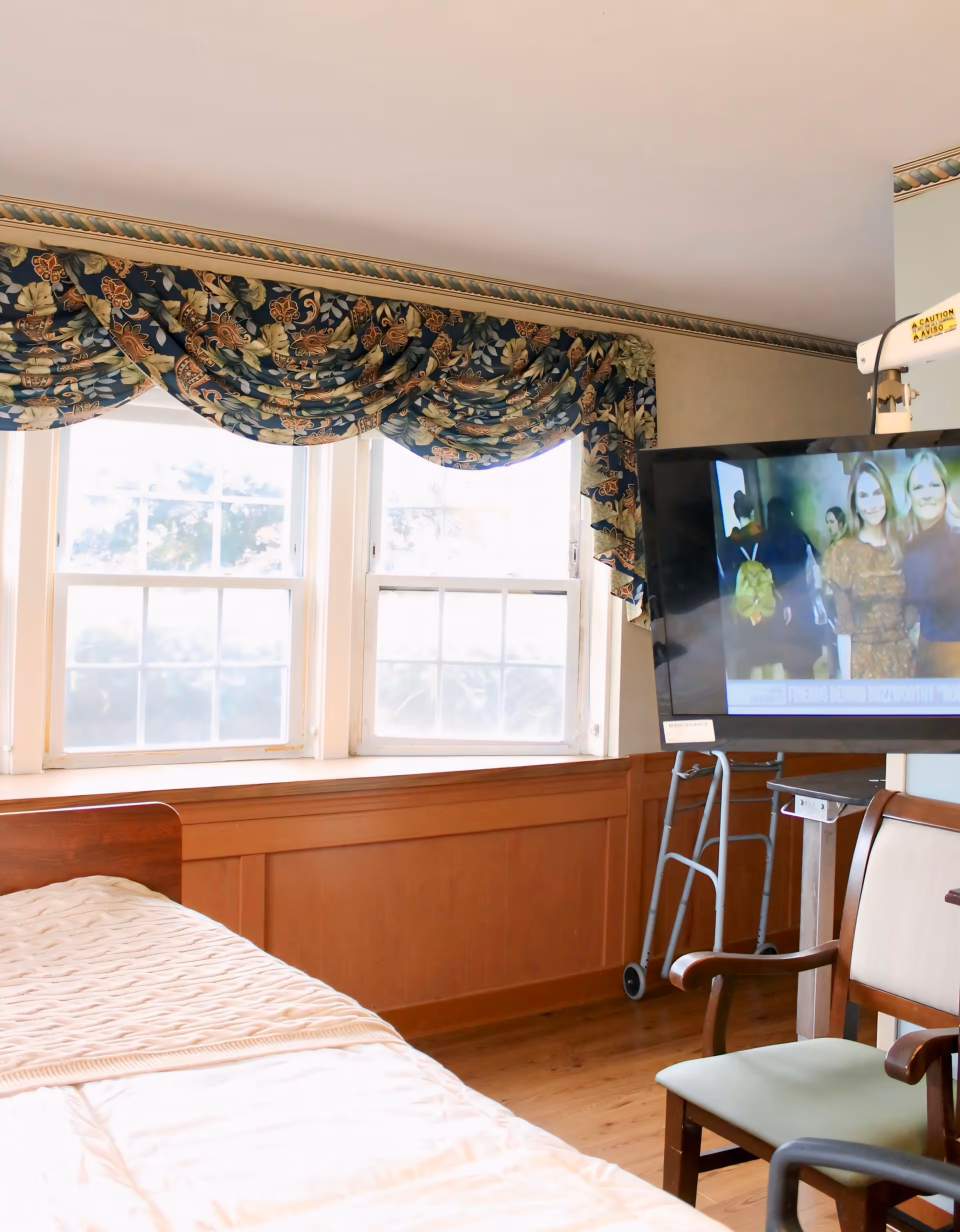 Nursing bedroom with a bed, floral valance curtains, and a mounted TV.