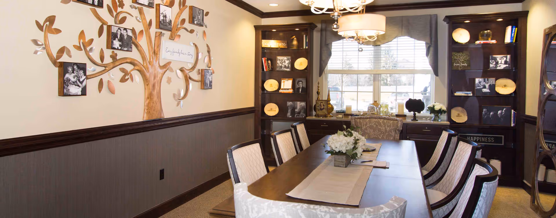 A well-lit dining room with a long wooden table surrounded by upholstered chairs. The table is decorated with a beige runner and a floral centerpiece. On the left wall, there is a decorative wooden tree with framed black and white family photos and a sign that reads 'Every family has a story.' The back wall features two dark wooden bookshelves filled with books, framed photos, decorative plates, and other ornaments. A large window with blinds and a valance lets in natural light, and a chandelier hangs above the table.