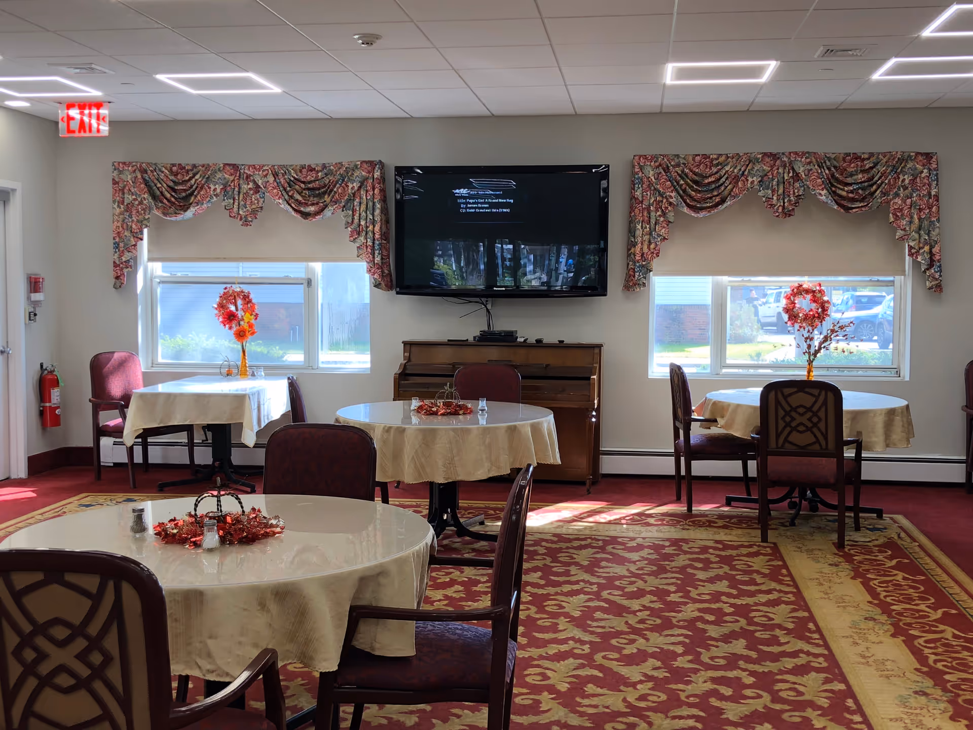 Decorated senior living dining room with round tables, chairs, a piano, and a TV.