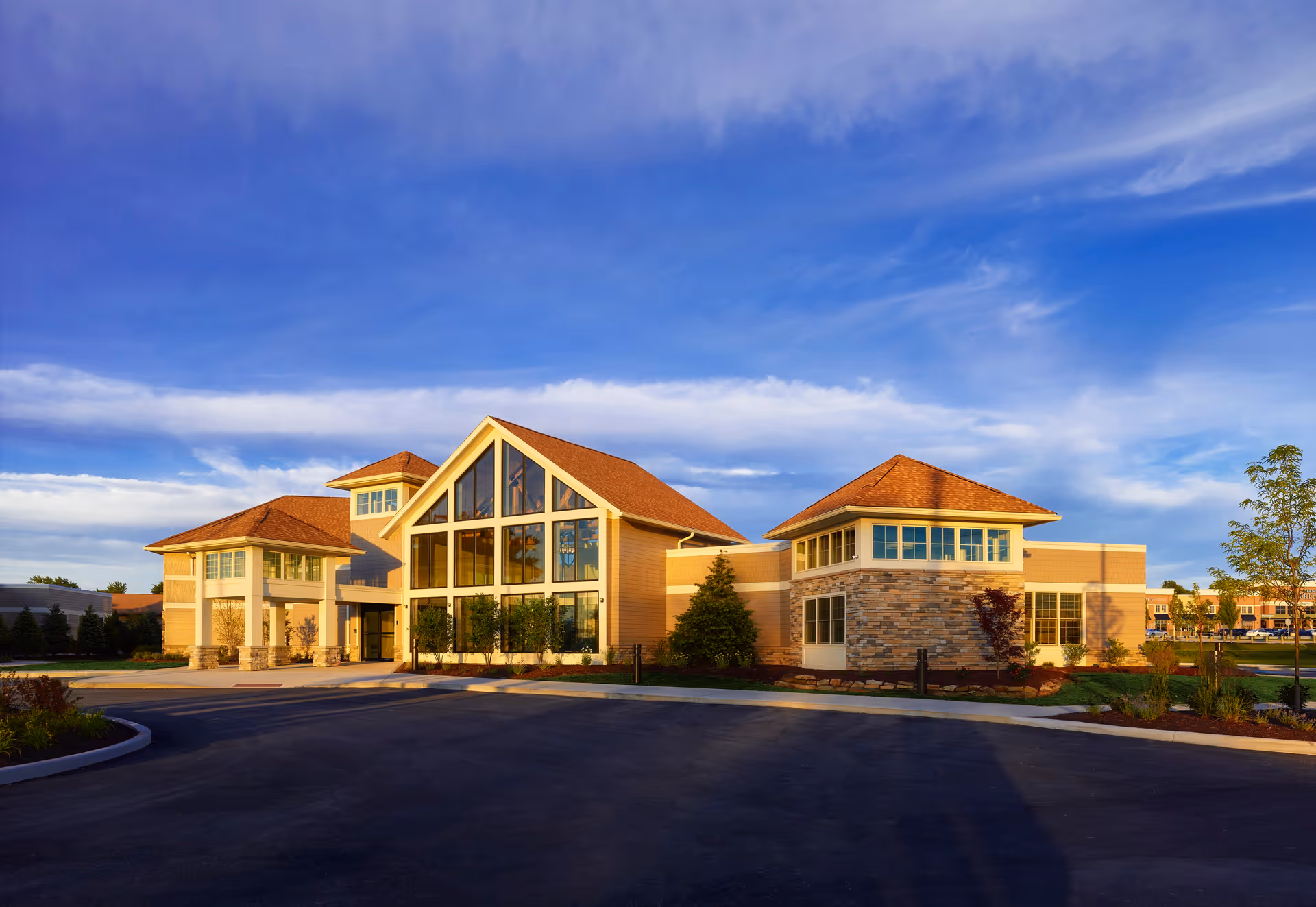 Modern senior living building with a large glass-front entrance, pitched roofs and landscaped grounds under a blue sky.