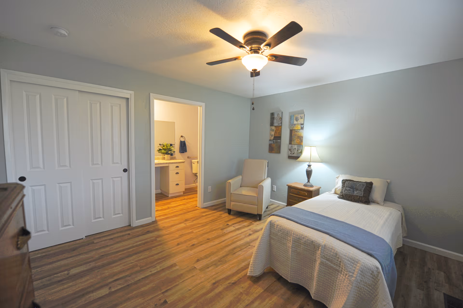 A cozy bedroom in an assisted living facility featuring a single bed with white bedding and a blue throw, a wooden nightstand with a lamp, a comfortable armchair, and a closet with white sliding doors. The room has light gray walls, wood flooring, and a ceiling fan with a light. An open door reveals a bathroom with a sink, mirror, and toilet.