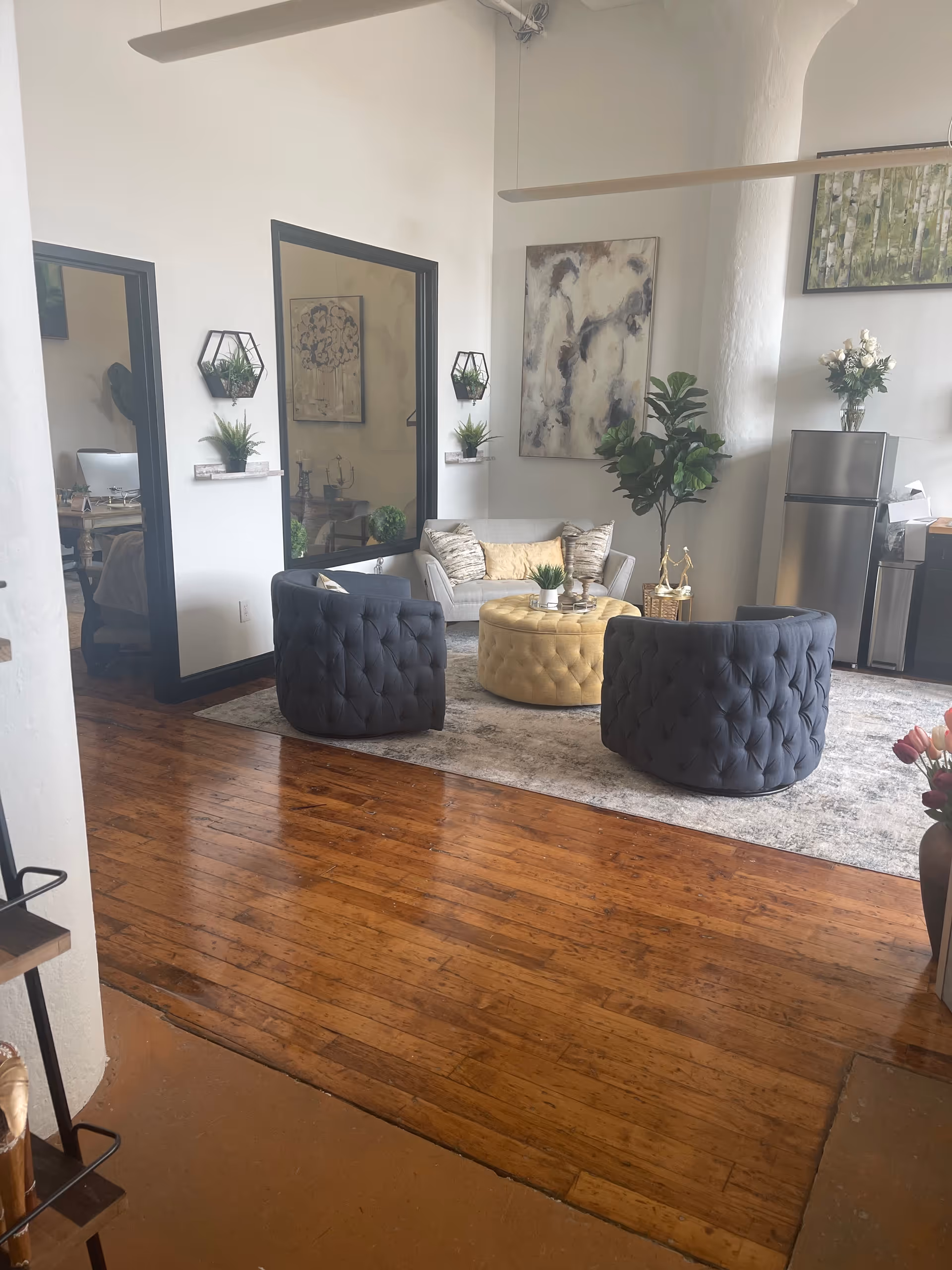 Bright modern living room with tufted ottomans, sofa, greenery, and a stainless fridge.