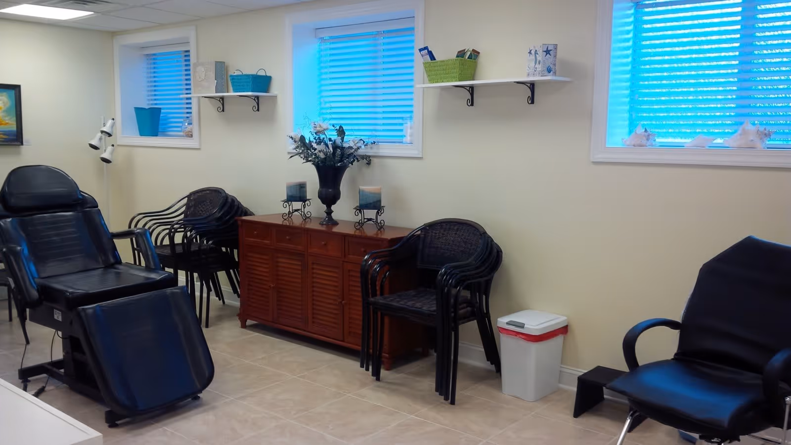 A clean and organized room with black reclining chairs, stacked black chairs, a wooden cabinet with decorative items including a vase with flowers and candles, two small windows with blue blinds, and a white trash bin with a red lid.