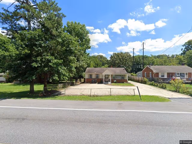 Small single-story brick building with a paved parking area and chain-link fence facing a road, flanked by trees and neighboring houses.