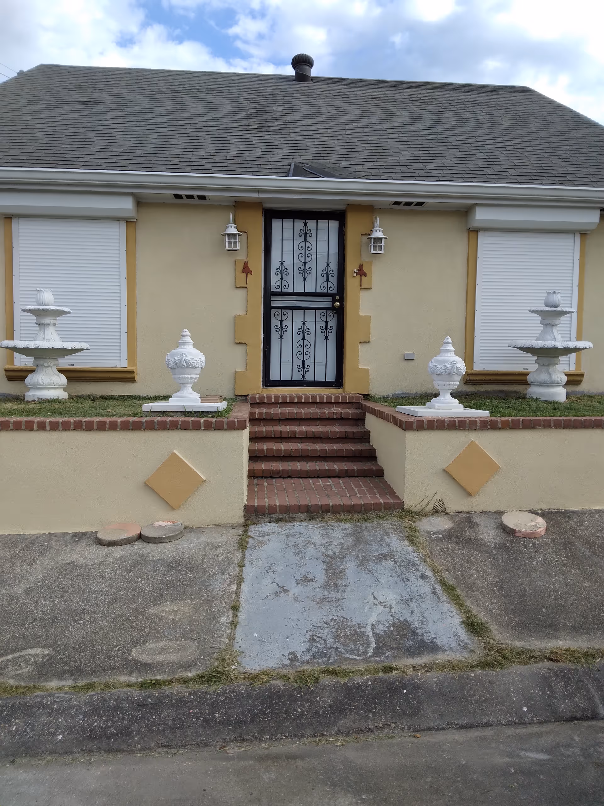 Front view of a beige house with brick steps, a wrought-iron door, and decorative fountains.