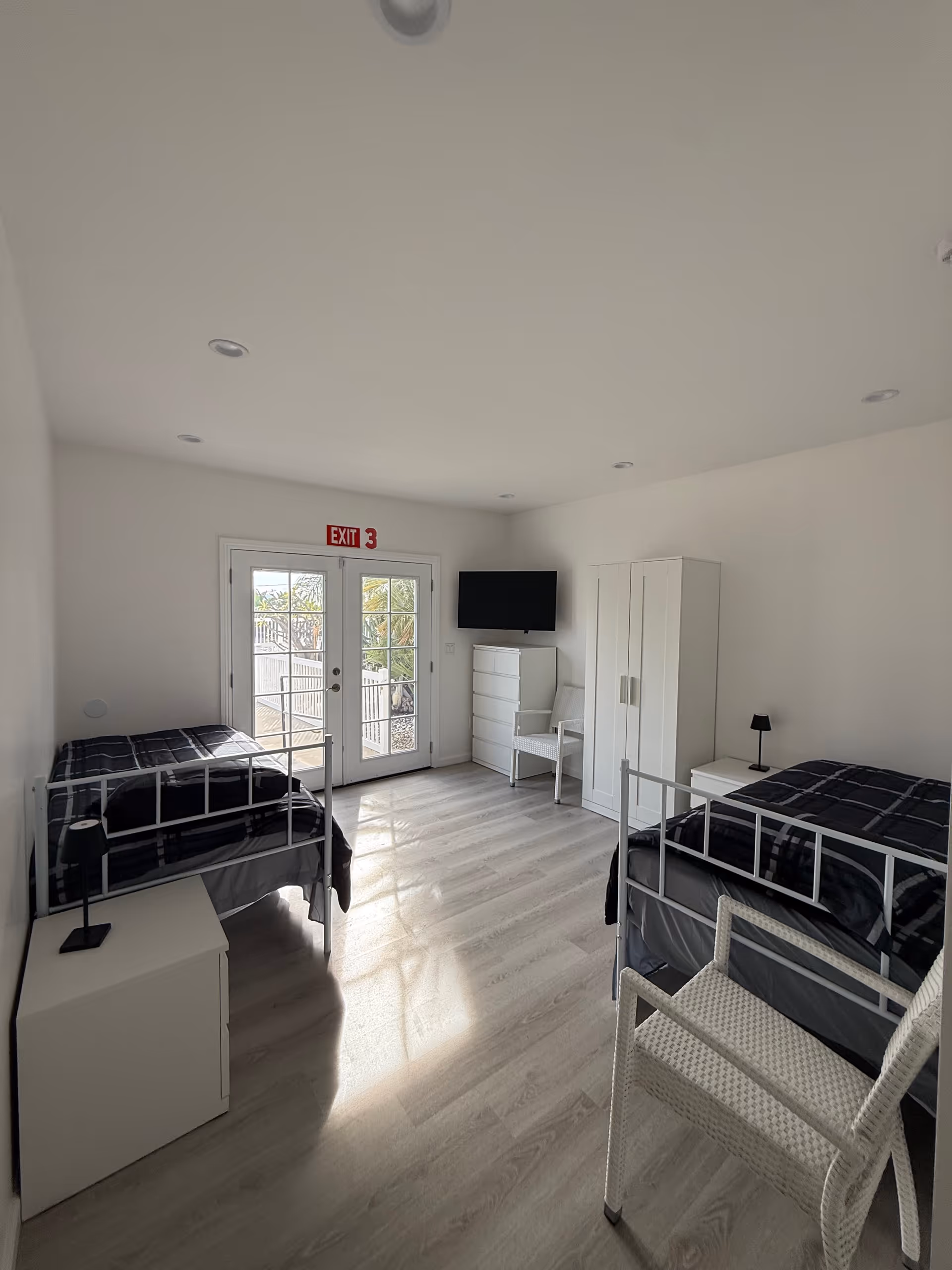 Bright twin-bedroom with two metal-framed beds, white furniture, dresser, wardrobe, and TV.