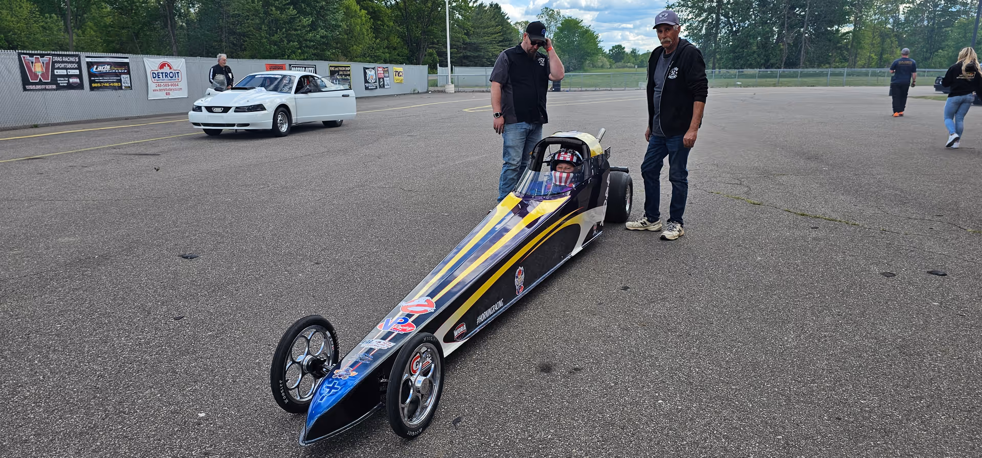 A long, slender dragster sits on a paved track as two men stand nearby.