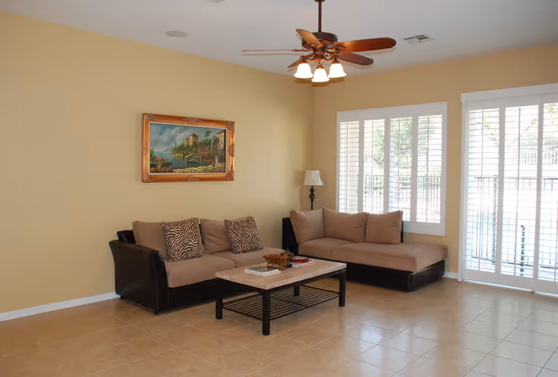 Living room with beige couches, coffee table, painting, ceiling fan, and large windows.