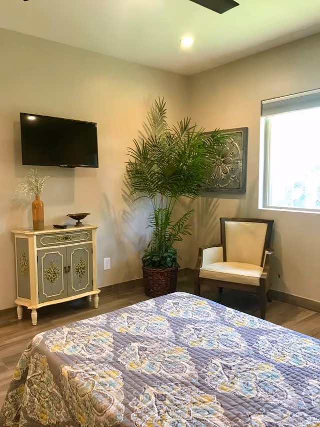 A cozy bedroom with a bed covered in a floral quilt, a vintage-style cabinet with decorative items on top, a wall-mounted flat screen TV, a large potted plant, an upholstered armchair, and a window letting in natural light.