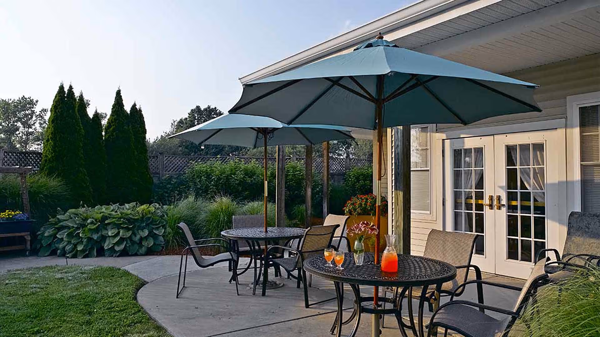 Outdoor patio area with round metal tables and chairs under large blue umbrellas. There is a pitcher and glasses with orange drink on one table, along with a small vase of flowers. The patio is adjacent to a building with white French doors and windows. Surrounding the patio are green bushes, tall trees, and a wooden fence.