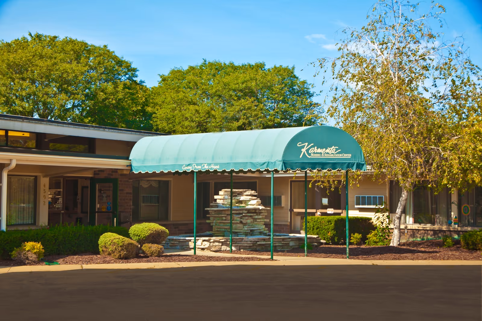 Exterior view of Karmenta Center entrance with green canopy, stone fountain, and surrounding landscaping.