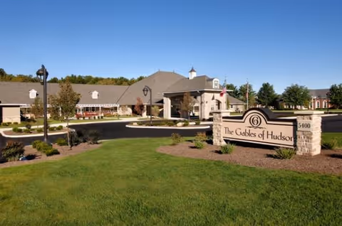 Front entrance and landscaped grounds of a senior living building with a sign reading "The Gables of Hudson" under a clear blue sky.