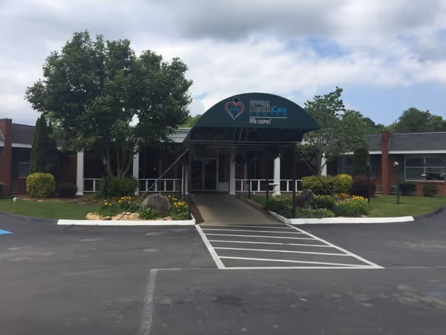 Front exterior view of Etowah Health Care Center with a covered entrance, surrounded by trees and landscaped greenery under a partly cloudy sky.