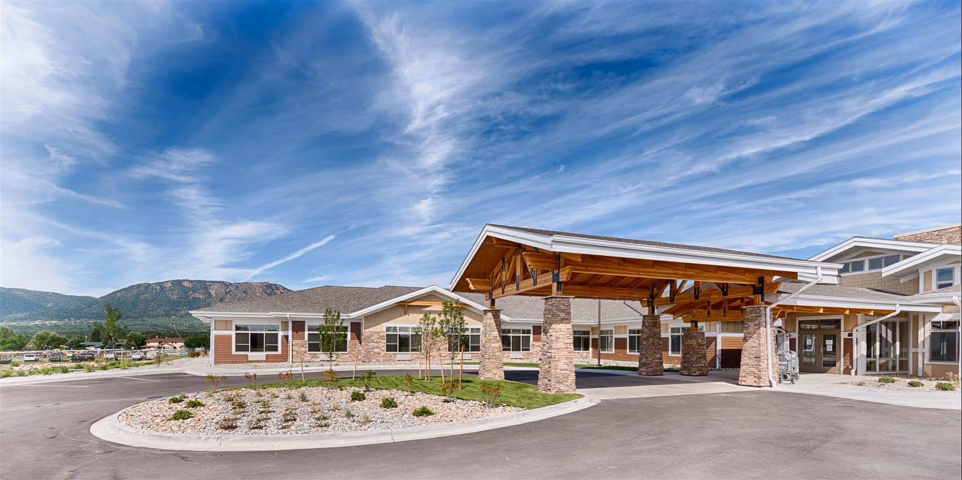 Exterior view of Bethesda Gardens Monument senior living facility showing a covered entrance with stone pillars, a driveway, landscaped area with small plants, and a mountainous background under a partly cloudy blue sky.