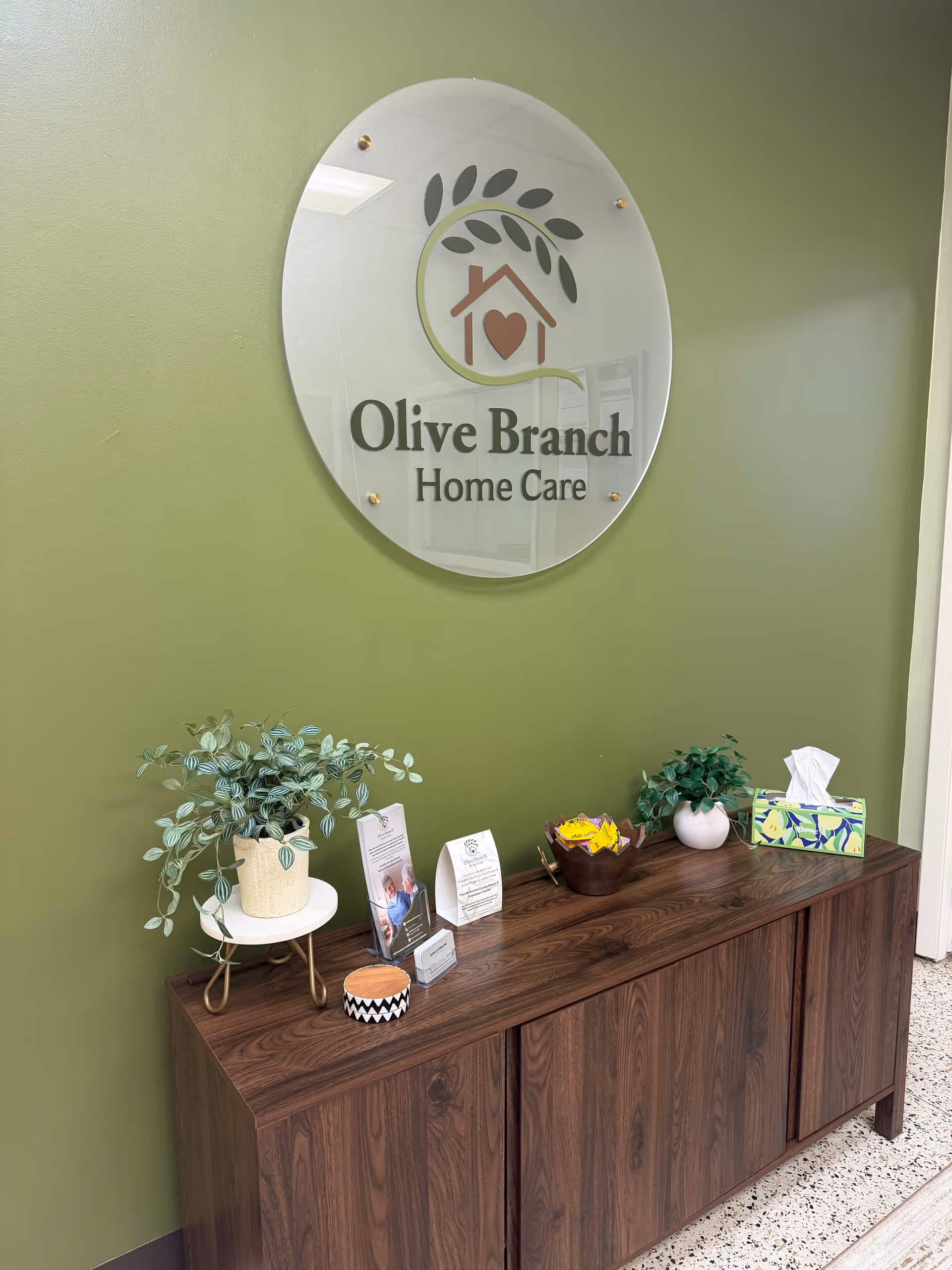 Interior lobby with Olive Branch Home Care sign and wooden cabinet with plants.