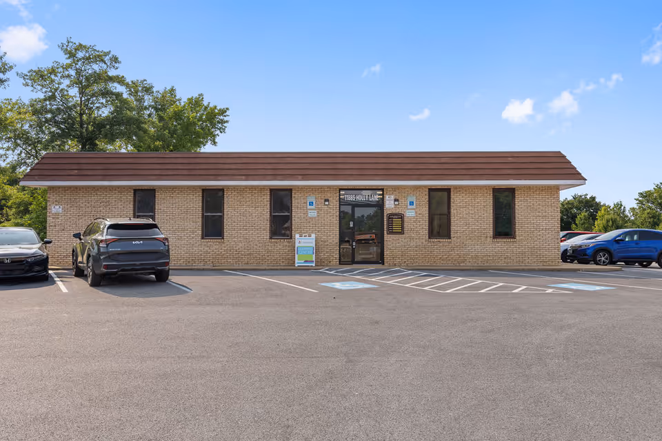 Brick facility facade with a parking lot and cars in front of the entrance.