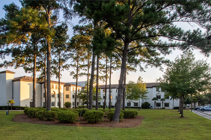 White three-story senior living building with a landscaped lawn and tall pine trees in front under a clear sky.