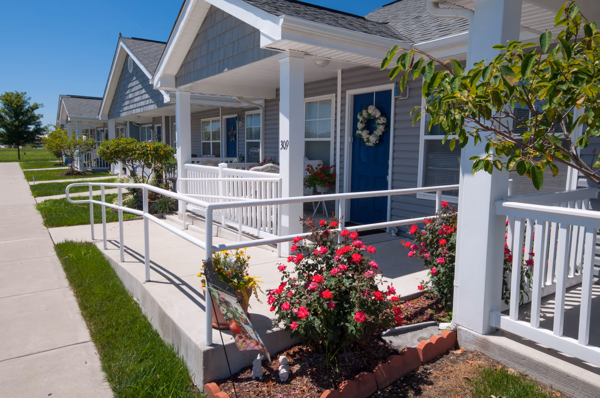 Exterior view of a senior living facility named Laurel Commons showing a row of single-story units with blue doors and white railings. There is a concrete walkway with a ramp, flower beds with red and yellow flowers, and small trees along the front porches under a clear blue sky.