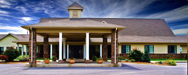 Front exterior view of a single-story building with a covered entrance supported by brick and white columns, surrounded by small bushes and plants under a partly cloudy sky.