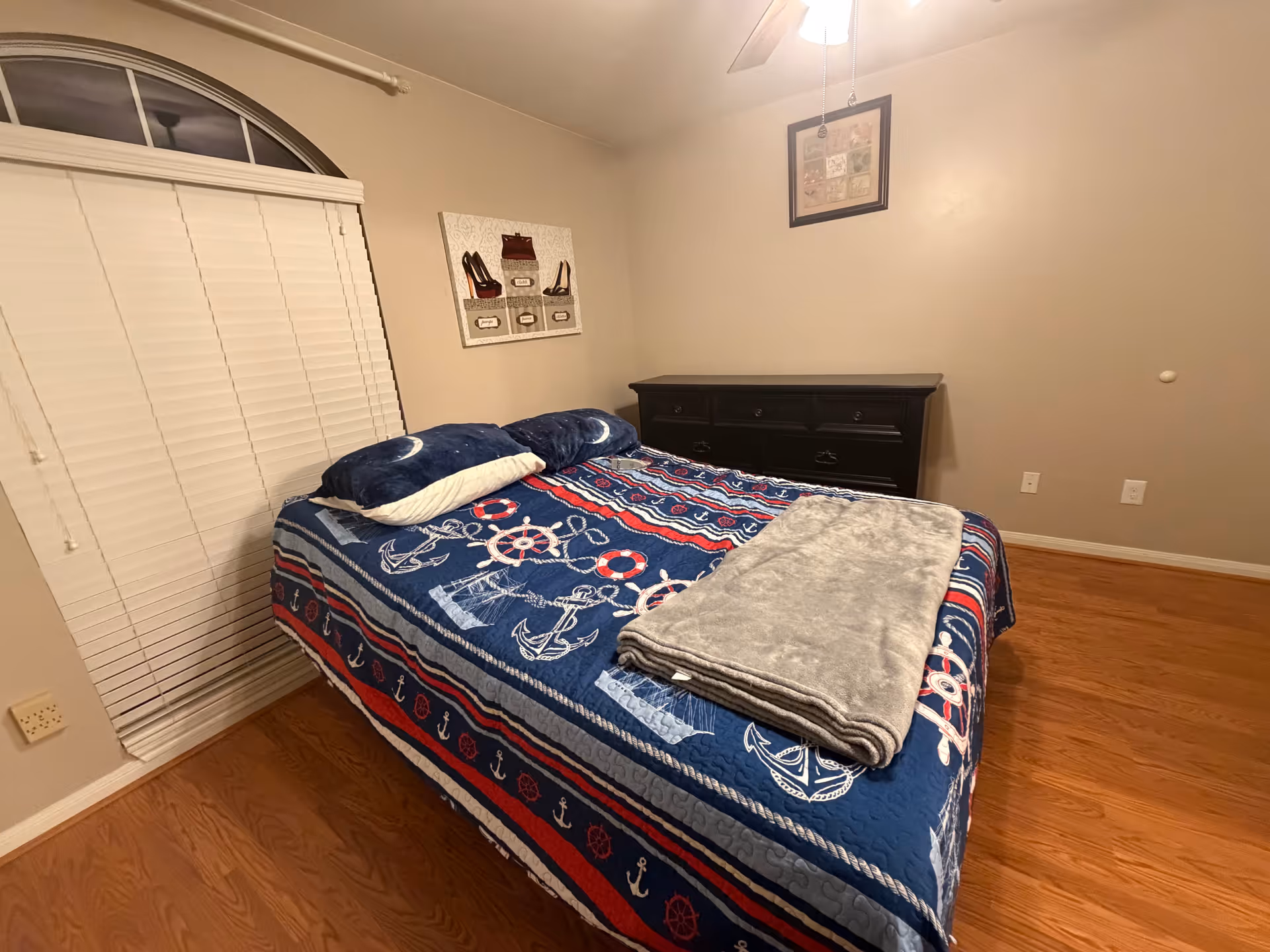 A small bedroom with nautical-themed bedspread, pillows, a dresser, and blinds.