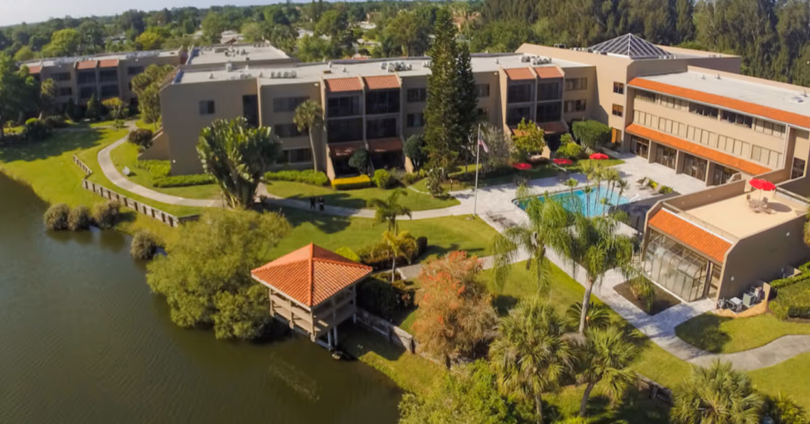 Aerial view of the Lakehouse West Retirement Estate showing multi-story buildings, a central courtyard with pool, a lakeside gazebo, and landscaped grounds.