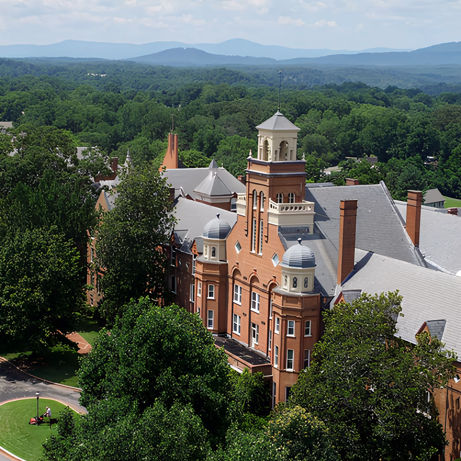 Aerial view of a large historic red-brick building with a central tower surrounded by trees and distant hills.
