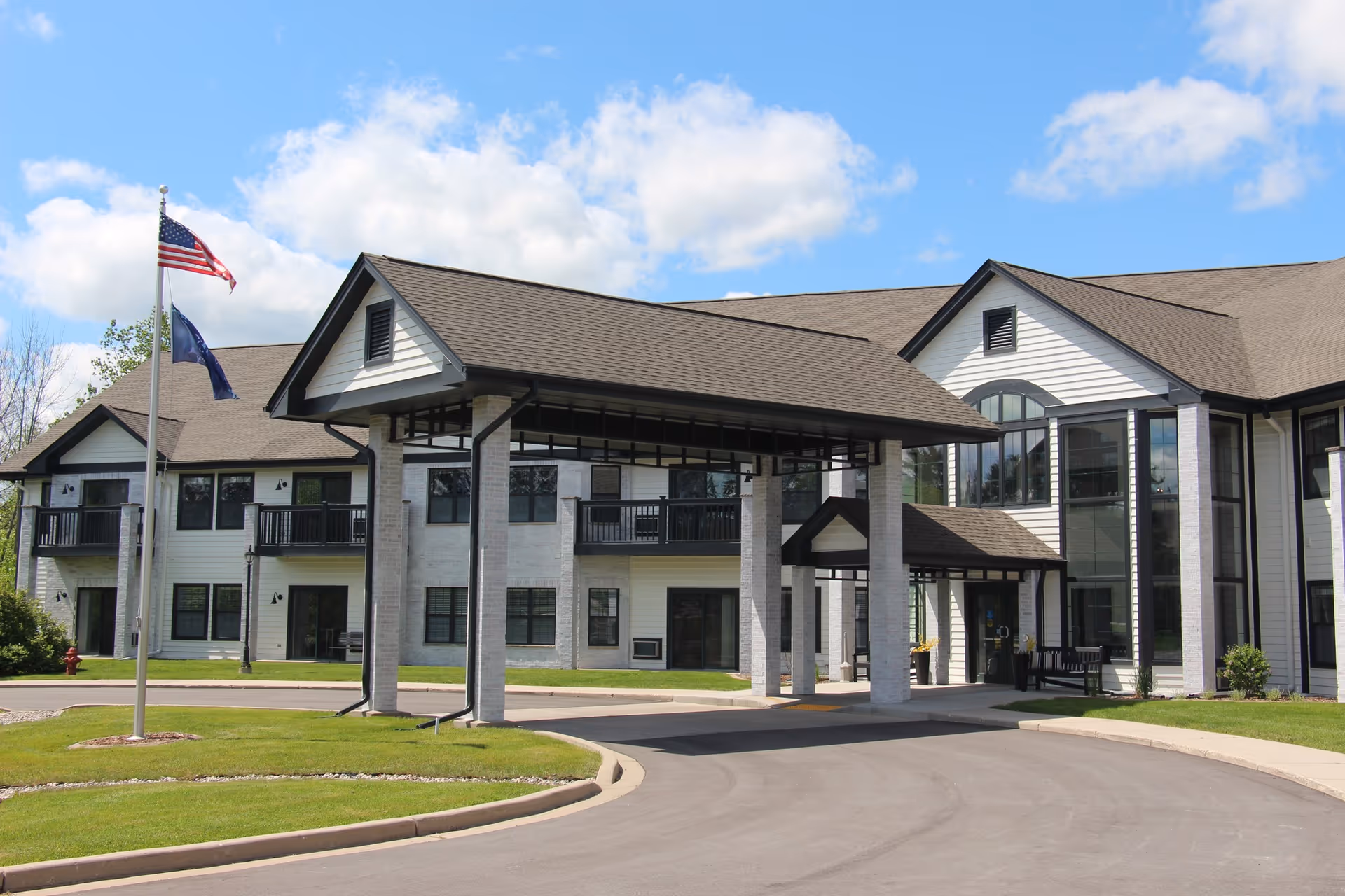 Front exterior of a two-story senior living facility with a covered entrance portico, flagpoles, and a landscaped driveway under a blue sky.