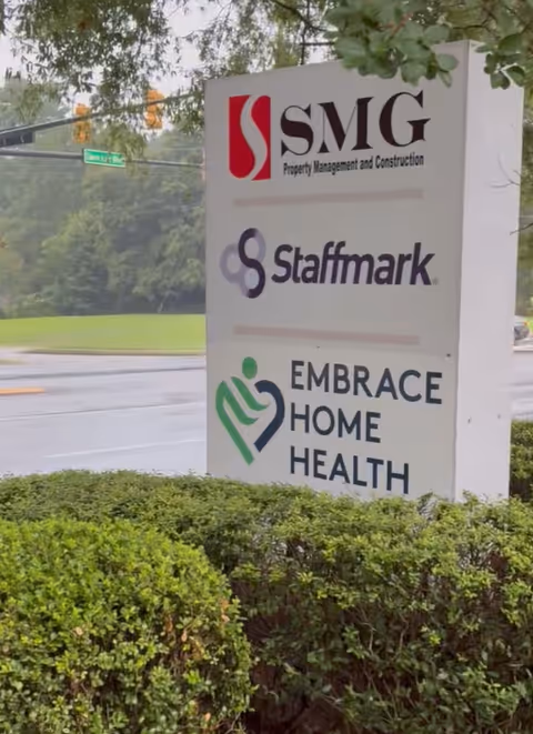 Outdoor sign displaying Embrace Home Health with SMG and Staffmark logos amid bushes.