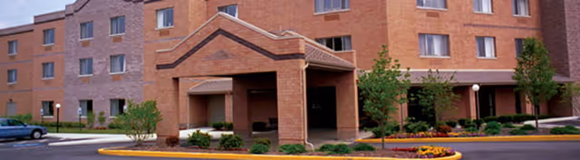 Front entrance of a multi-story brick building with a covered porte-cochere, landscaped beds, and a driveway.