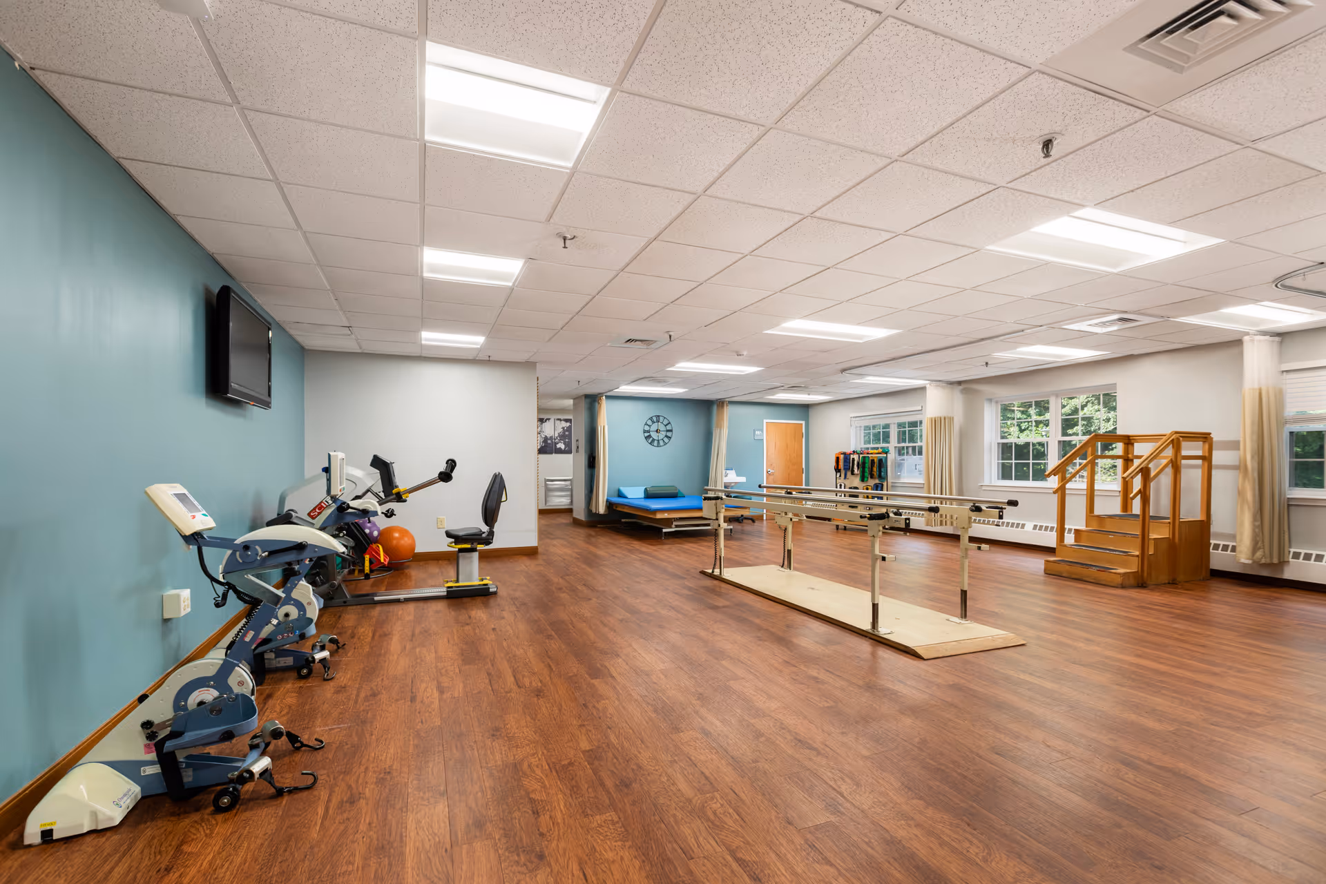 Interior rehabilitation gym at Kimberly Hall North with exercise machines, parallel bars, and wooden flooring.