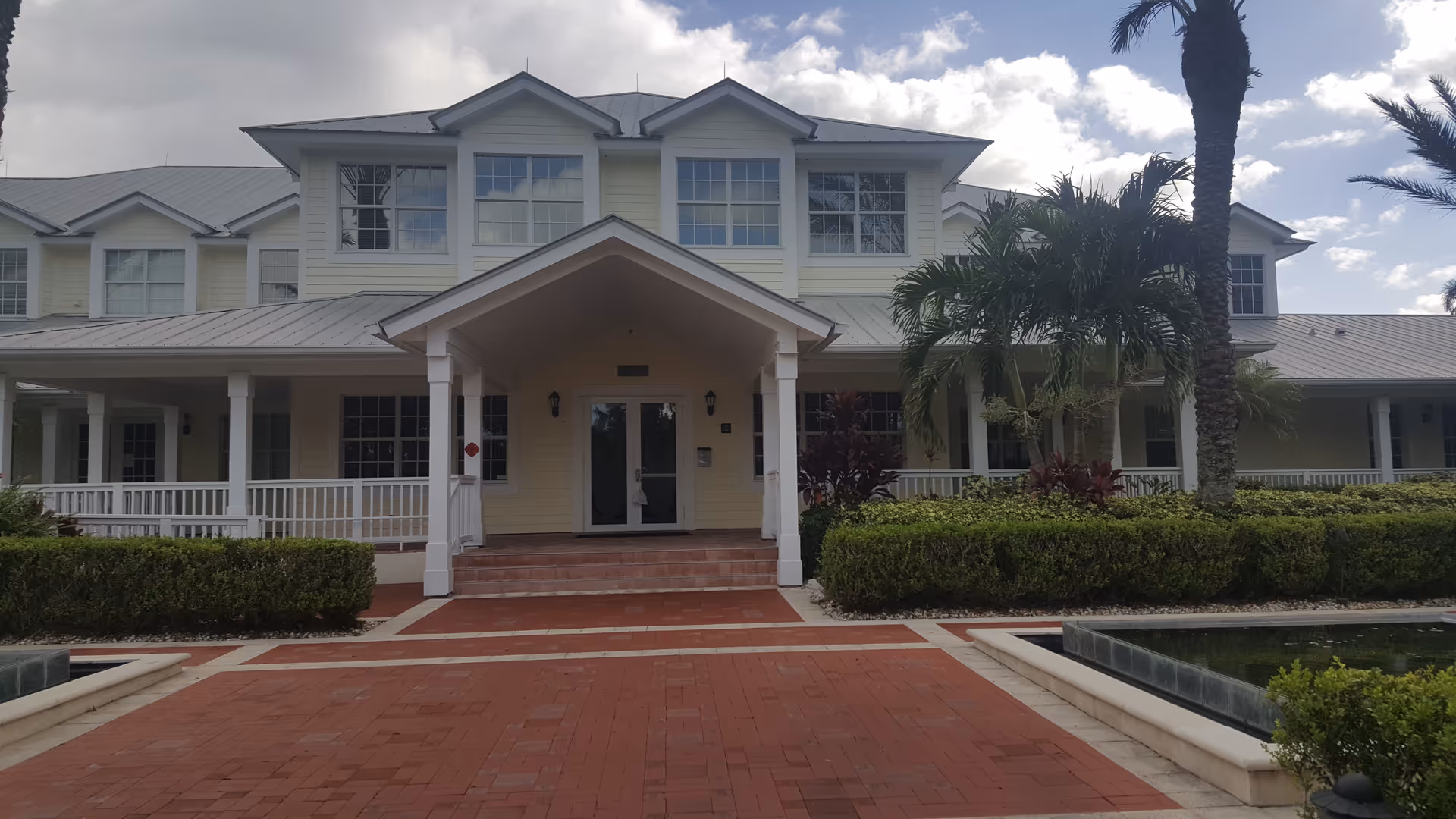 Two-story light-colored senior living facility with brick walkway, entrance portico, and palm trees.