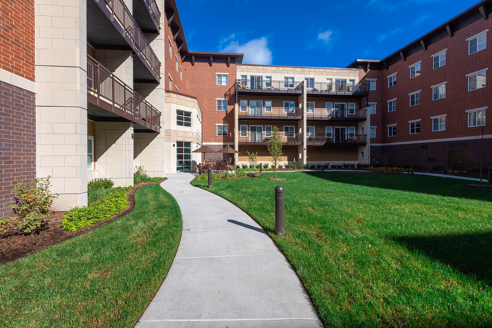 Outdoor courtyard area of The Carrington at Lincolnwood facility with a curved concrete walkway, green grass, small trees, and shrubs. The building surrounding the courtyard is made of brick and light-colored stone with balconies and windows. The sky is clear and blue with a few clouds.
