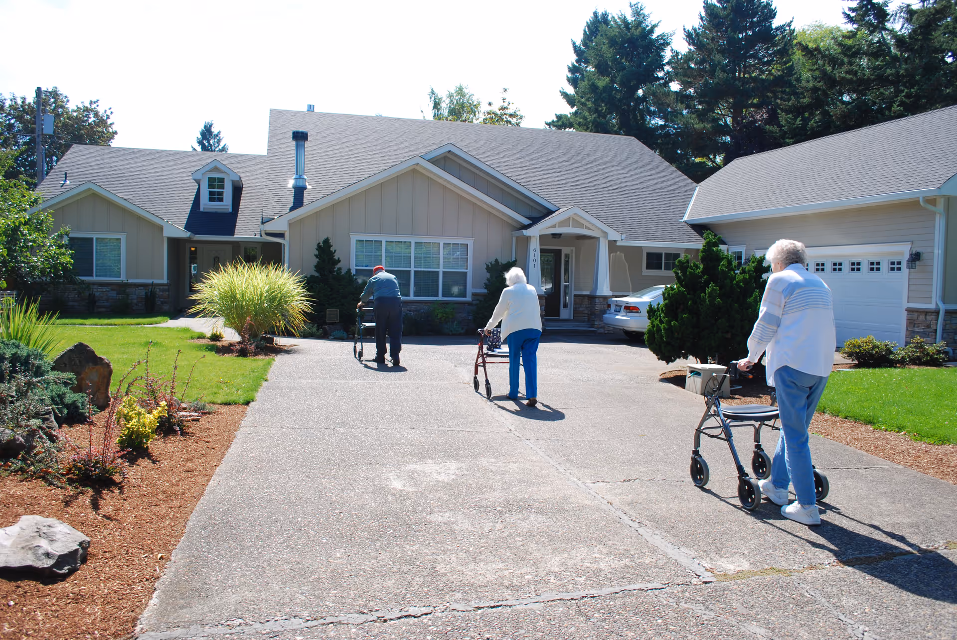 Three elderly residents using walkers walk toward the front entrance of a single-story senior living home.