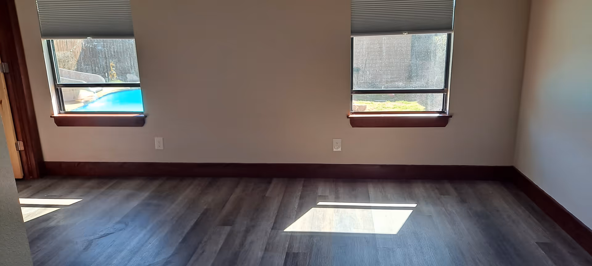 An empty room with wooden flooring, two windows with blinds, and baseboard trim.