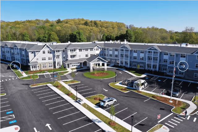 Aerial view of The Woods Retirement Community showing the building's front entrance, circular driveway, and large parking lot with trees in the background.