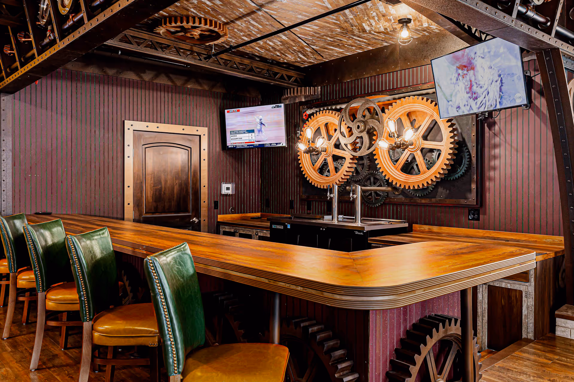 Interior view of a bar area with a wooden counter and green leather bar stools. The walls are decorated with large wooden gears and two mounted flat-screen TVs. The ceiling has exposed metal beams and a single hanging light bulb.