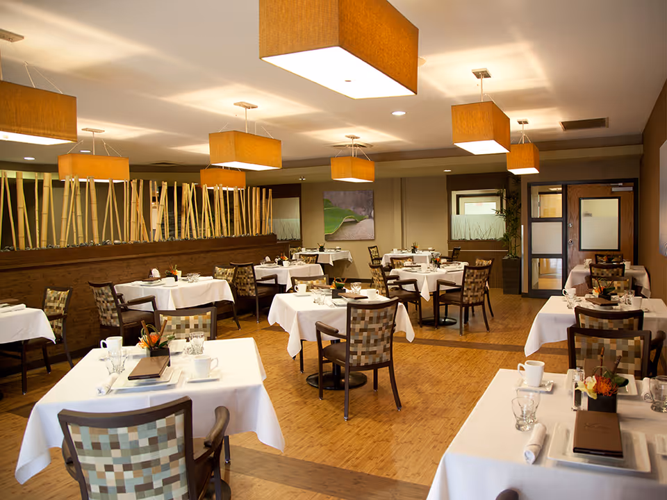 A well-lit dining room with multiple tables covered in white tablecloths, each set with cups, glasses, napkins, and small floral centerpieces. The room features wooden chairs with patterned upholstery, modern square pendant lights hanging from the ceiling, and a decorative wooden divider with vertical bamboo sticks. There are paintings on the walls and a door with frosted glass panels in the background.