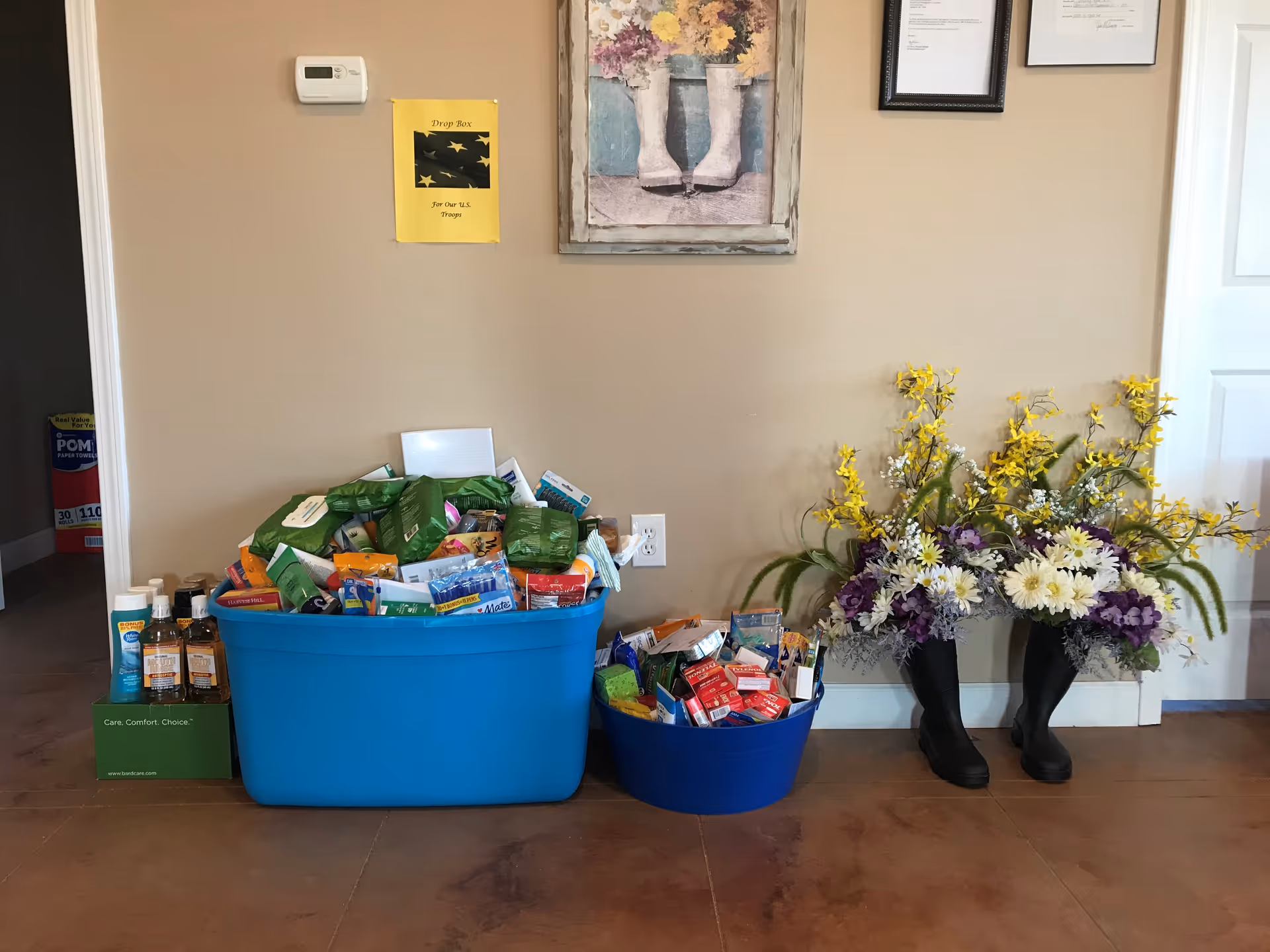 Beige interior hall with blue bins overflowing with packaged items and a tall floral arrangement.