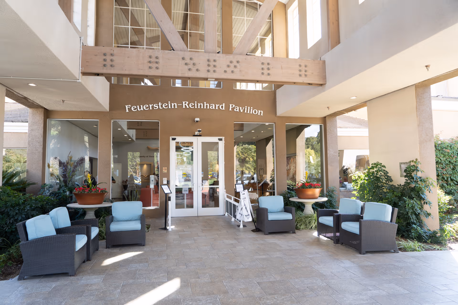 Entrance to the Feuerstein-Reinhard Pavilion at Heritage Pointe featuring glass double doors, large windows, and a covered seating area with cushioned wicker chairs and potted plants on either side.