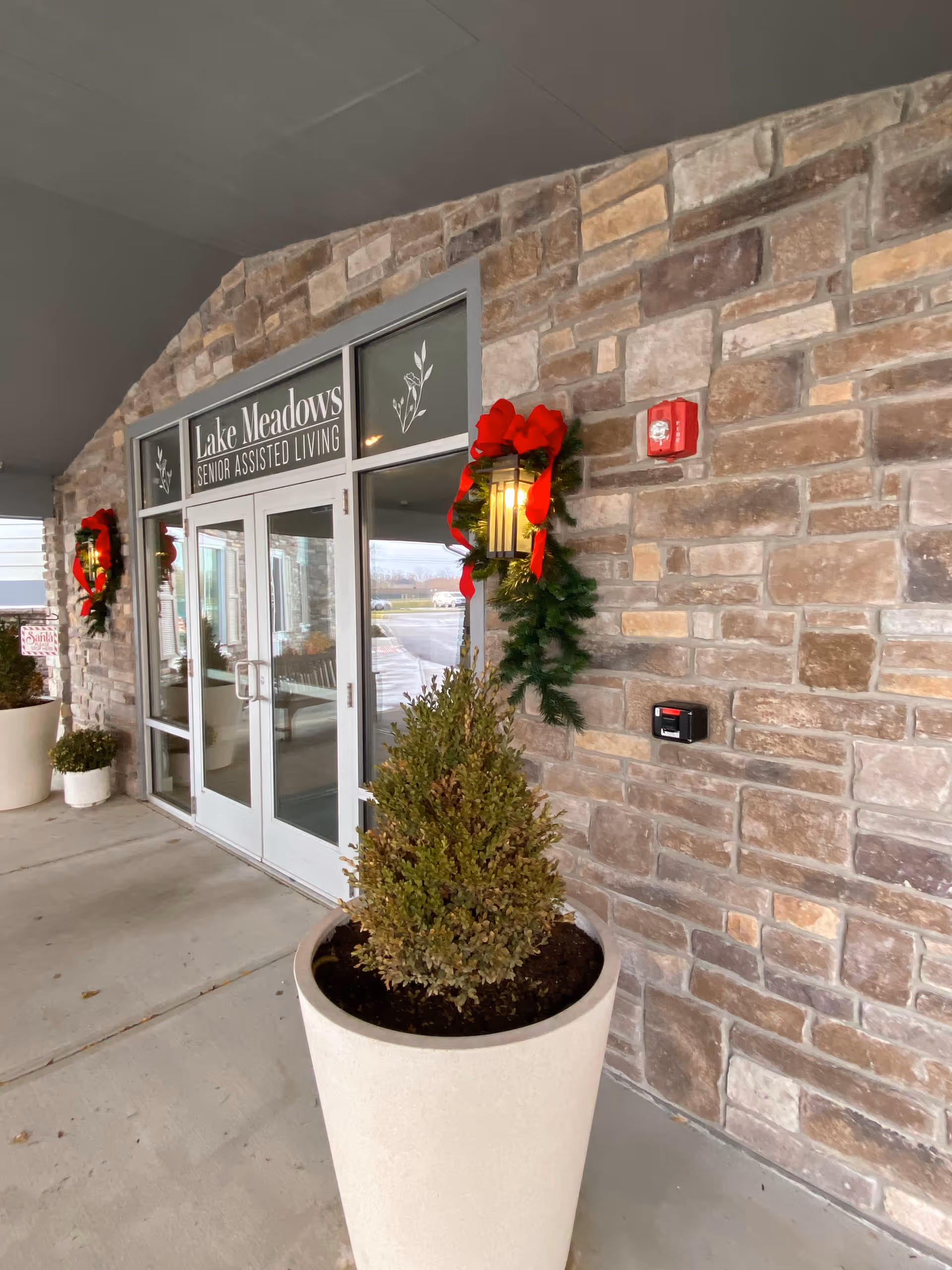 Entrance of Lake Meadows Senior Assisted Living with glass double doors, stone facade, potted plants and holiday wreaths.