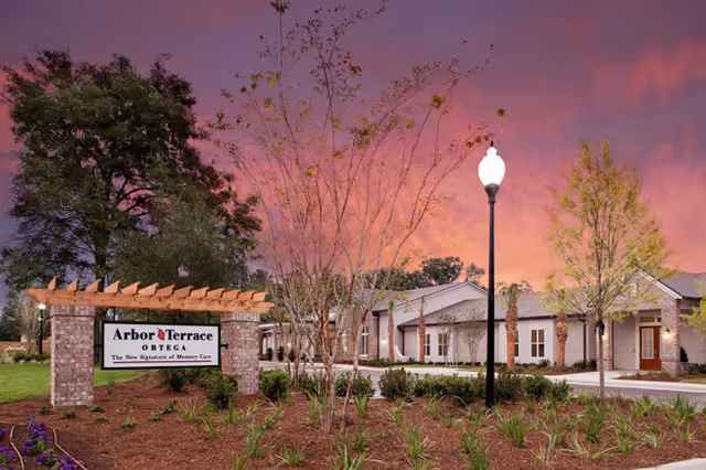 Entrance sign for Arbor Terrace Ortega in landscaped grounds in front of a single-story senior living building under a pink sunset sky.