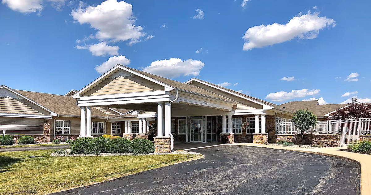 Front entrance of a single-story senior living facility with a covered porte-cochère supported by white columns, landscaped shrubs, and a paved driveway under a blue sky.