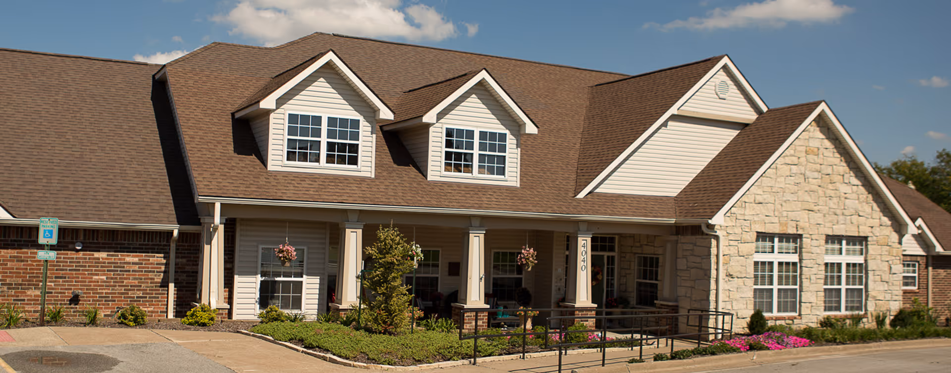Front exterior of a single-story residential-style building with a covered entrance, dormer windows, brick and stone facade, and landscaped beds.