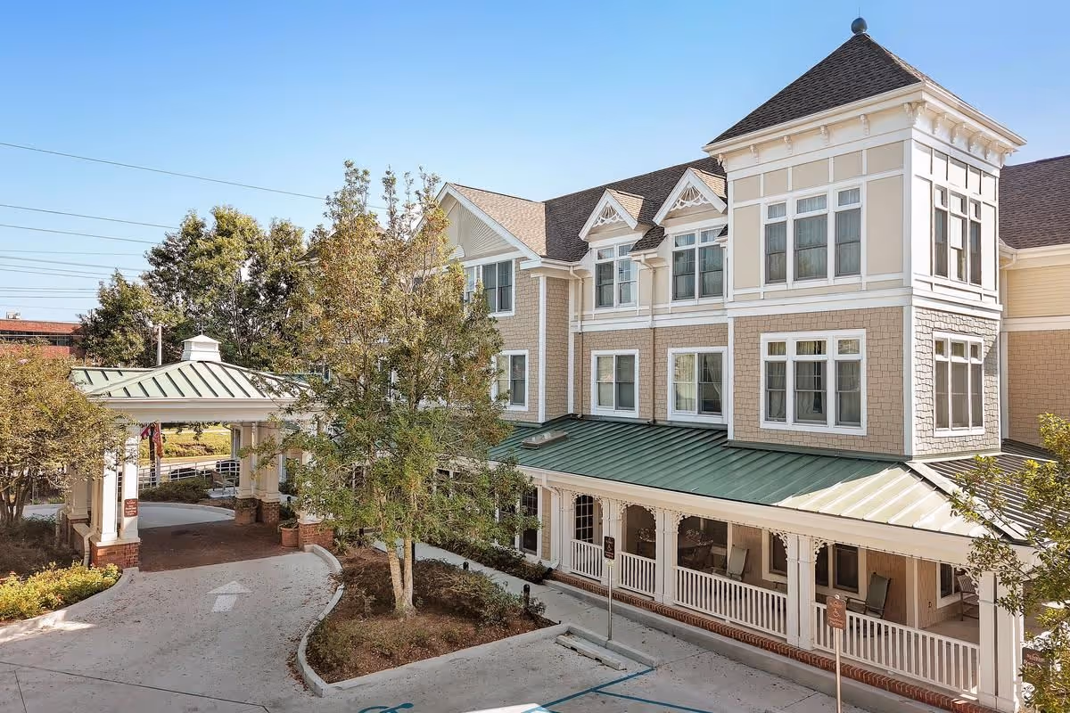 Exterior view of a multi-story senior living facility building with beige siding and white trim, featuring a covered entrance with a green metal roof, a porch with rocking chairs, and surrounding trees and landscaping under a clear blue sky.