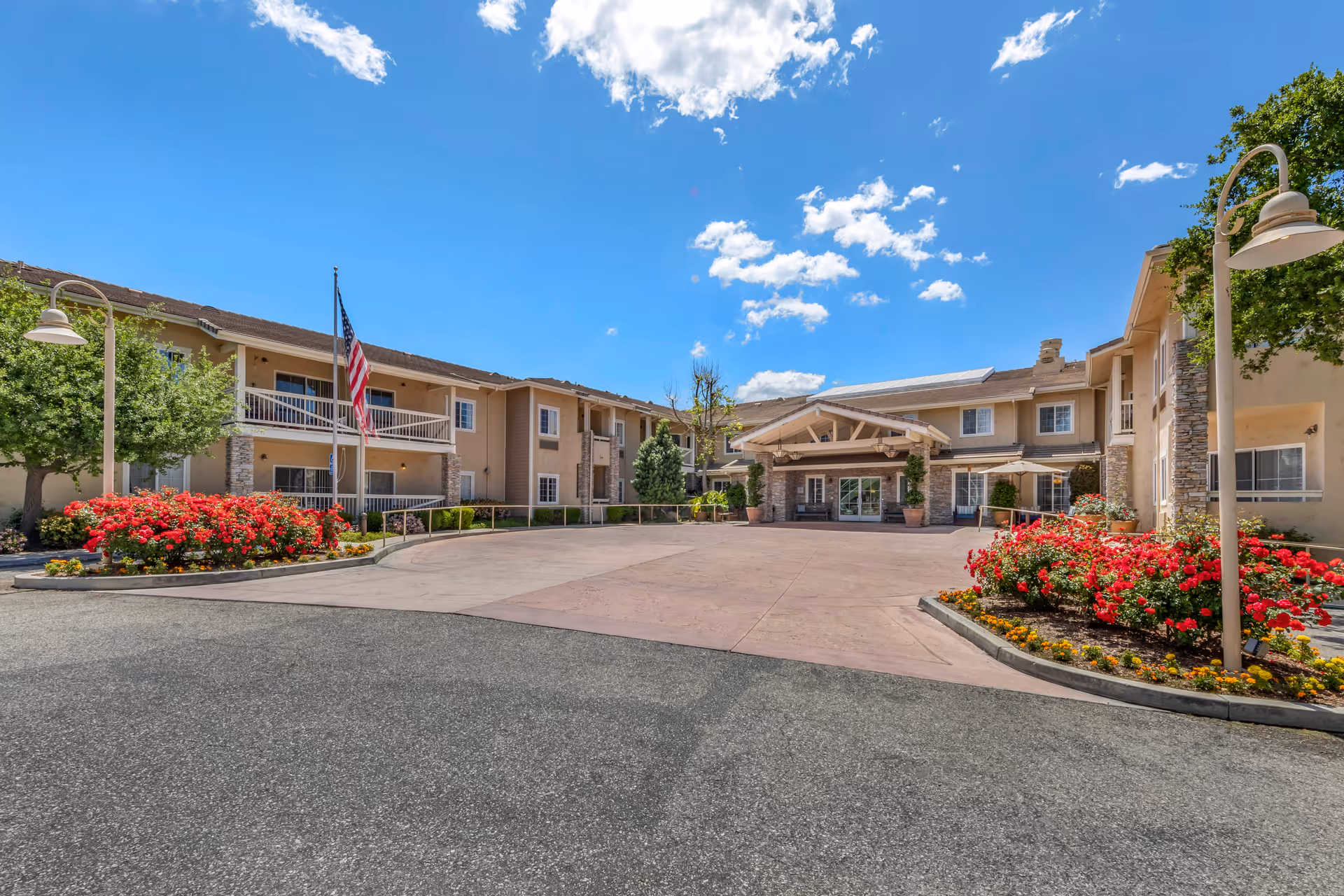 Front exterior view of Meadowbrook of Agoura Hills senior living facility with a clear blue sky, an American flag, well-maintained flower beds with red flowers, and a paved driveway leading to the entrance.