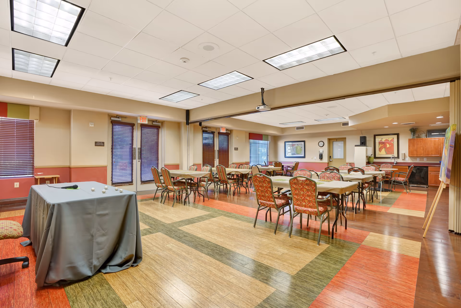 A spacious dining room with multiple tables and chairs arranged neatly. The room has colorful patterned flooring, beige walls with a red accent, and large windows with blinds. There is a covered table in the foreground and a kitchen area with cabinets in the background. The ceiling has recessed lighting and a projector mounted.
