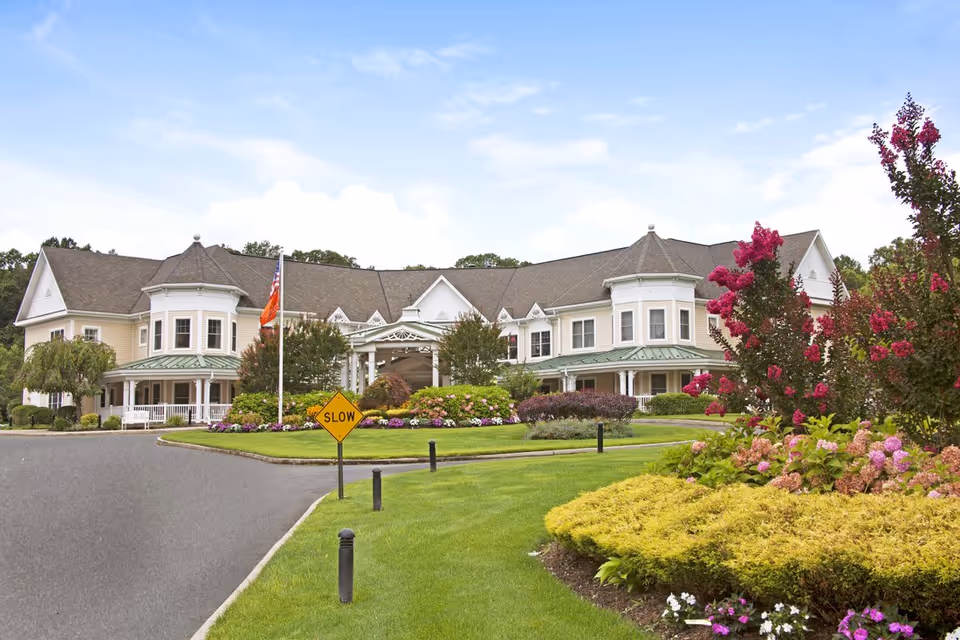 Exterior view of a large, elegant senior living facility building with a well-maintained garden and colorful flowers in the foreground. The building has a light-colored facade, multiple windows, and a covered entrance. A yellow 'SLOW' traffic sign is visible near the driveway.